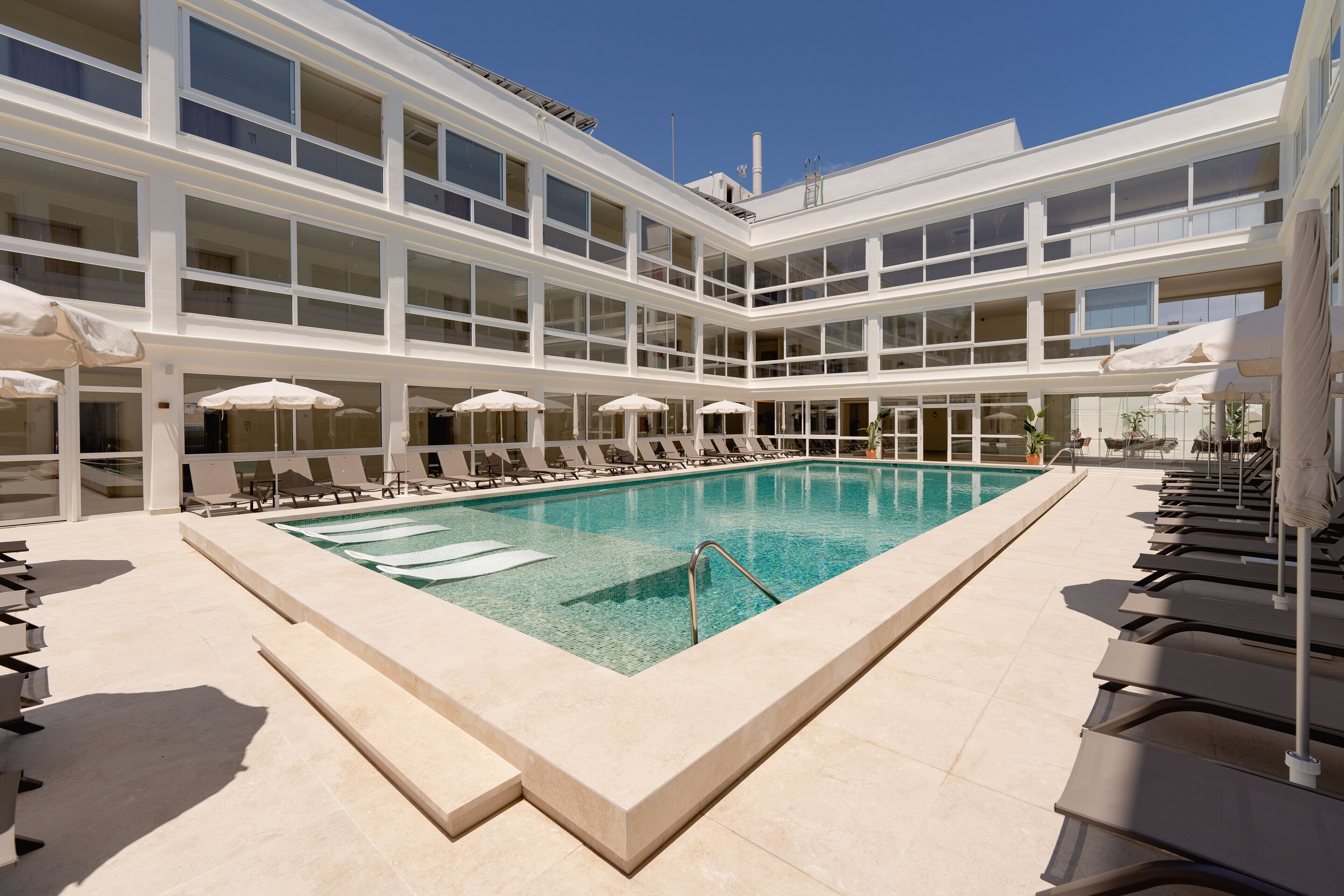 Un hotel moderno con una amplia piscina cubierta, varias zonas de descanso con sombrillas y un cielo azul despejado.
