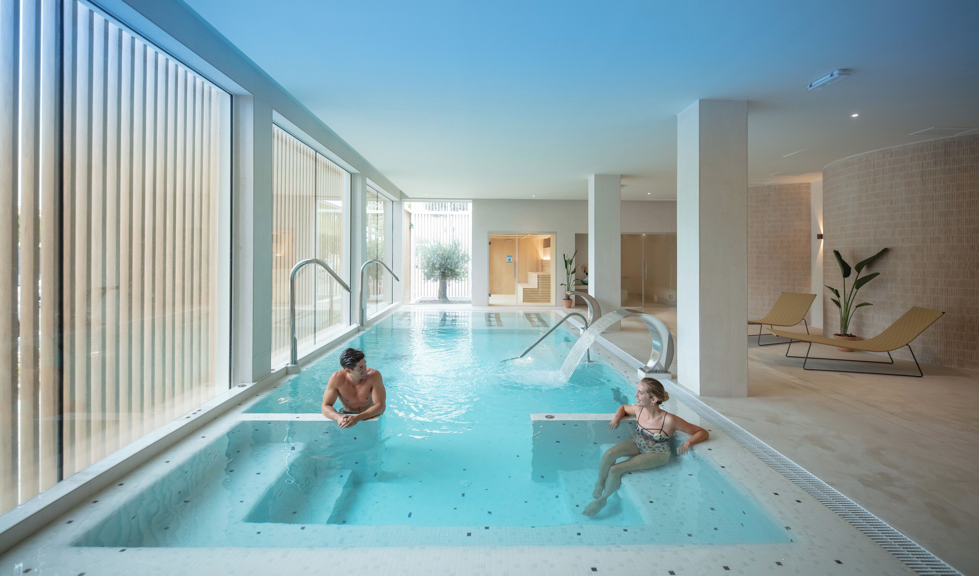 An indoor swimming pool with clear water, a tiled floor, and a glass partition on one side.