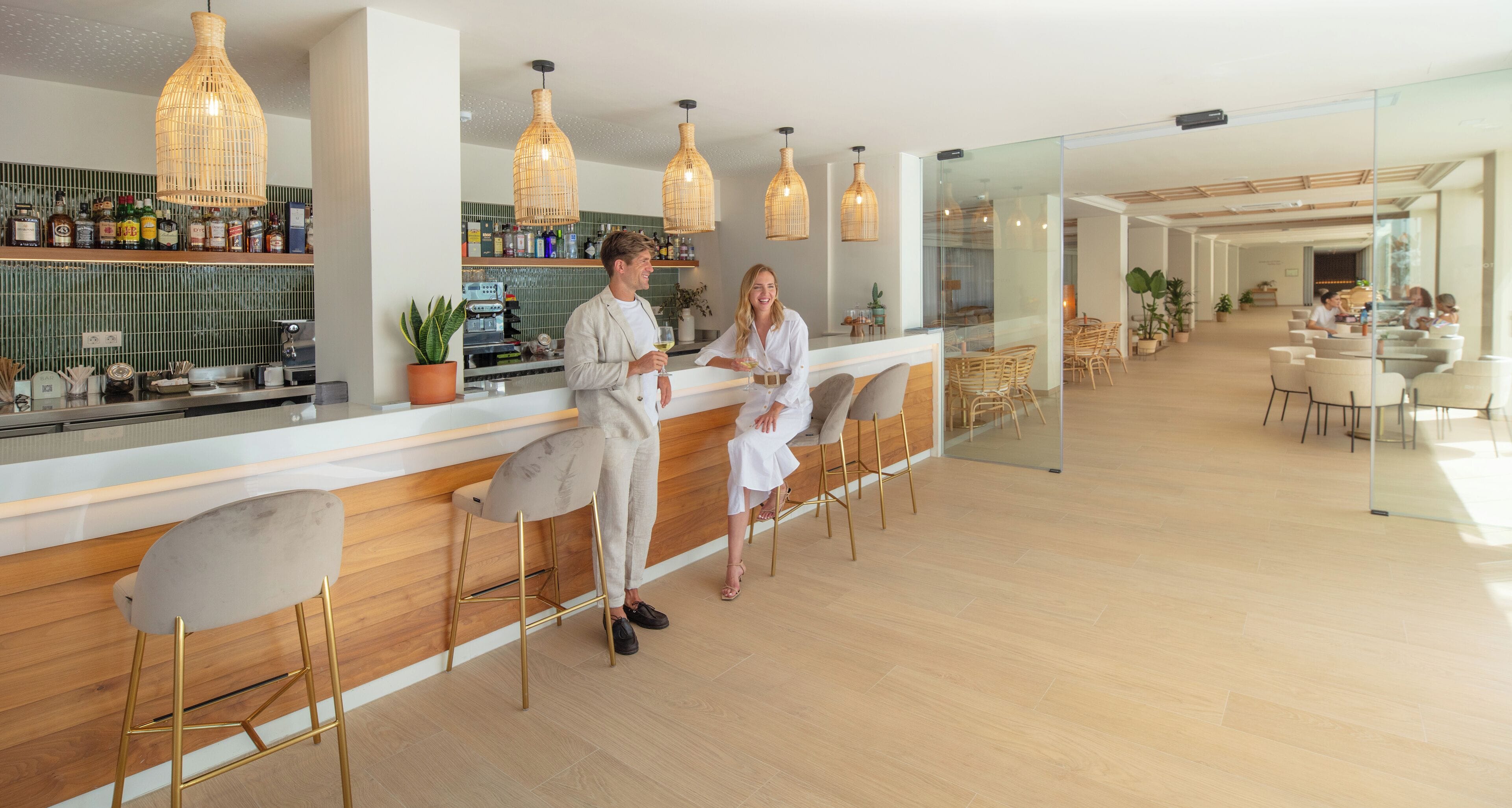 Un moderno bar con mostradores de madera, lámparas colgantes de mimbre y un separador de vidrio que lo distingue del área de comedor.