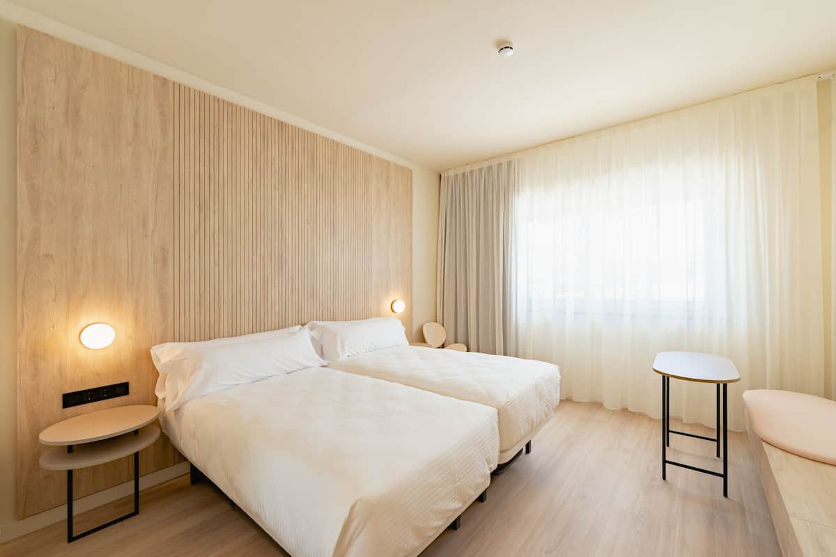 A hotel room with two beds, a round table, a chair, and a large window with curtains.