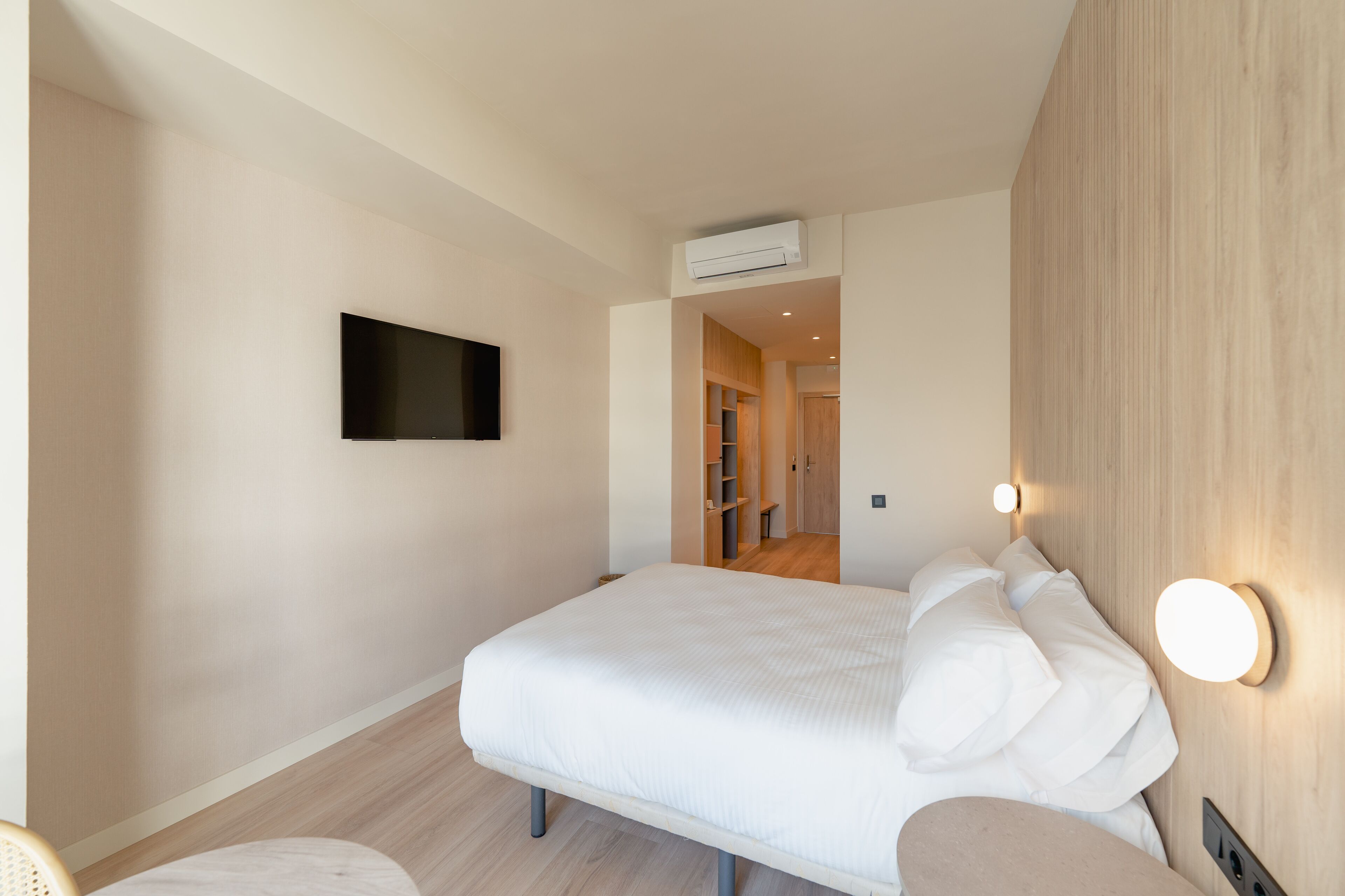 A hotel room with a bed, a flat-screen TV mounted on the wall, a bedside table, and a wall-mounted light fixture.