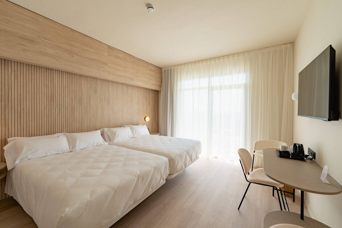 A hotel room with two beds, a desk, a chair, a television, and a window with curtains.