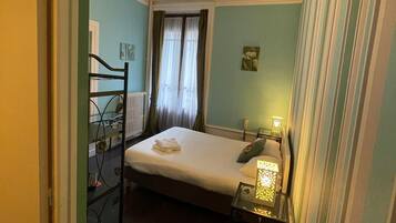 A hotel room with a bed, a desk with a lamp, a radiator, and a window with curtains.