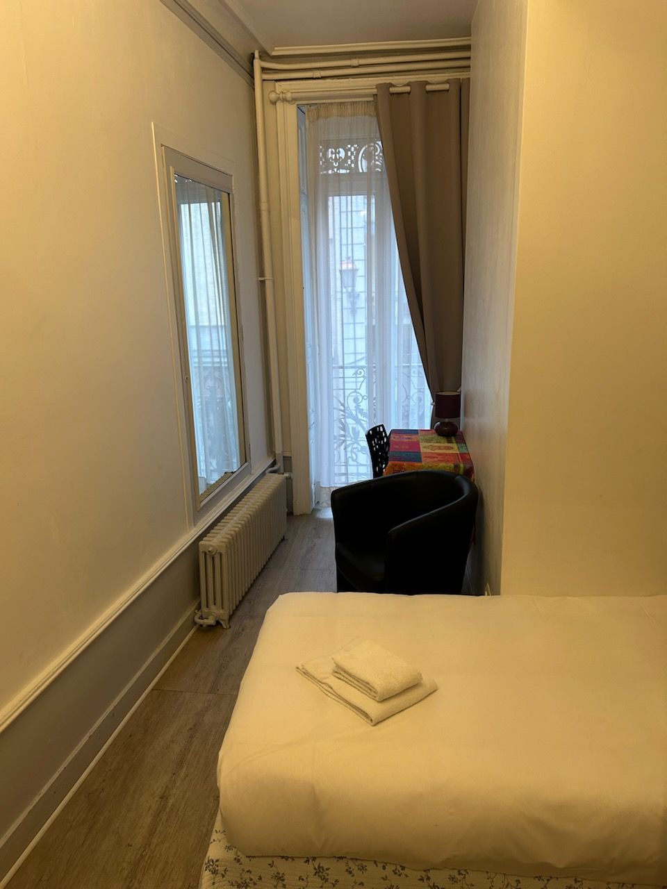 A hotel room with a bed, a radiator, a window with curtains, and a chair.
