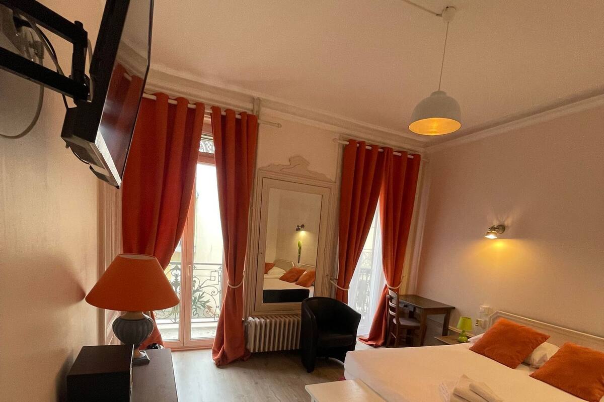 A hotel room with a bed, a desk with a lamp, a chair, a small table, and a window with curtains.