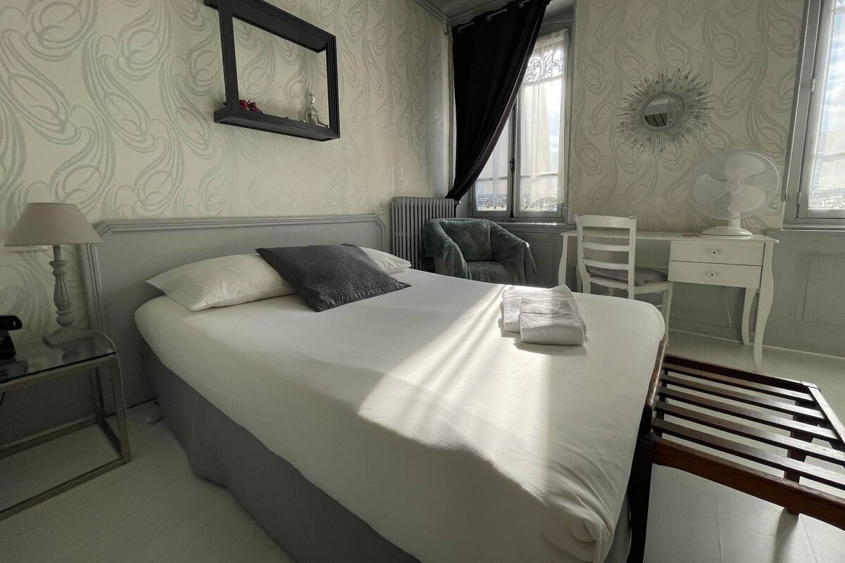 A neatly made bed with pillows, a bedside table with a lamp, a chair, a desk, and a radiator.