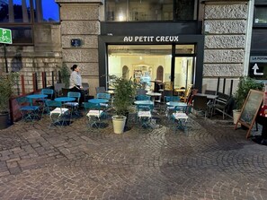 An outdoor café area with blue chairs and tables, a sign reading "AU PETIT CREUX," and a cobblestone pavement. - Authentic by Balladins, Le Carré d'Aix (Aix-les-Bains)