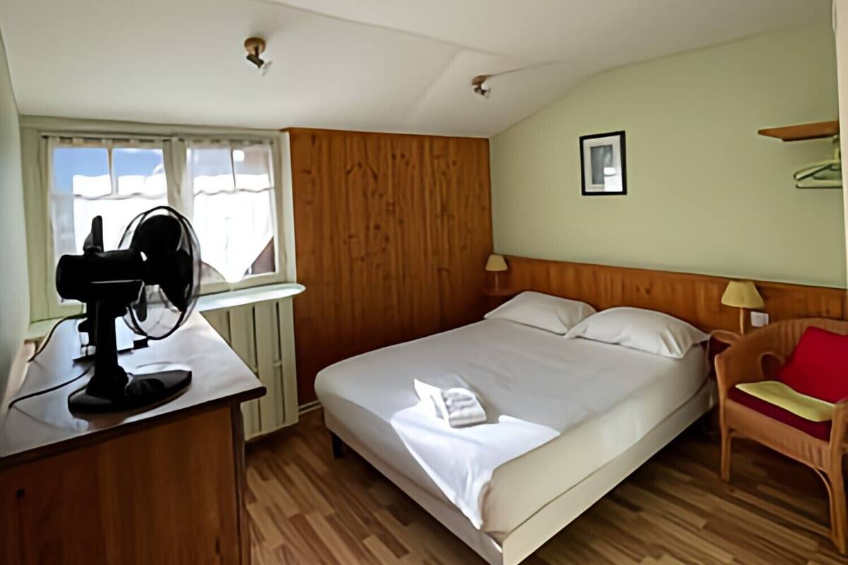 A bedroom with a bed, a desk with a fan, a chair, and a window with curtains.