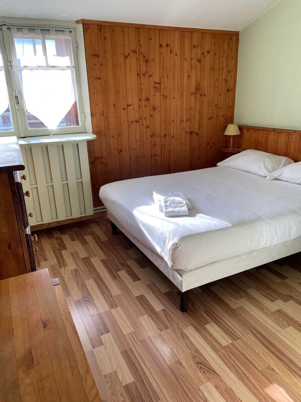 A bedroom with a wooden floor, a bed with white linens, a window with white curtains, and a small bedside table with a lamp. - Authentic by Balladins, Le Carré d'Aix (Aix-les-Bains)