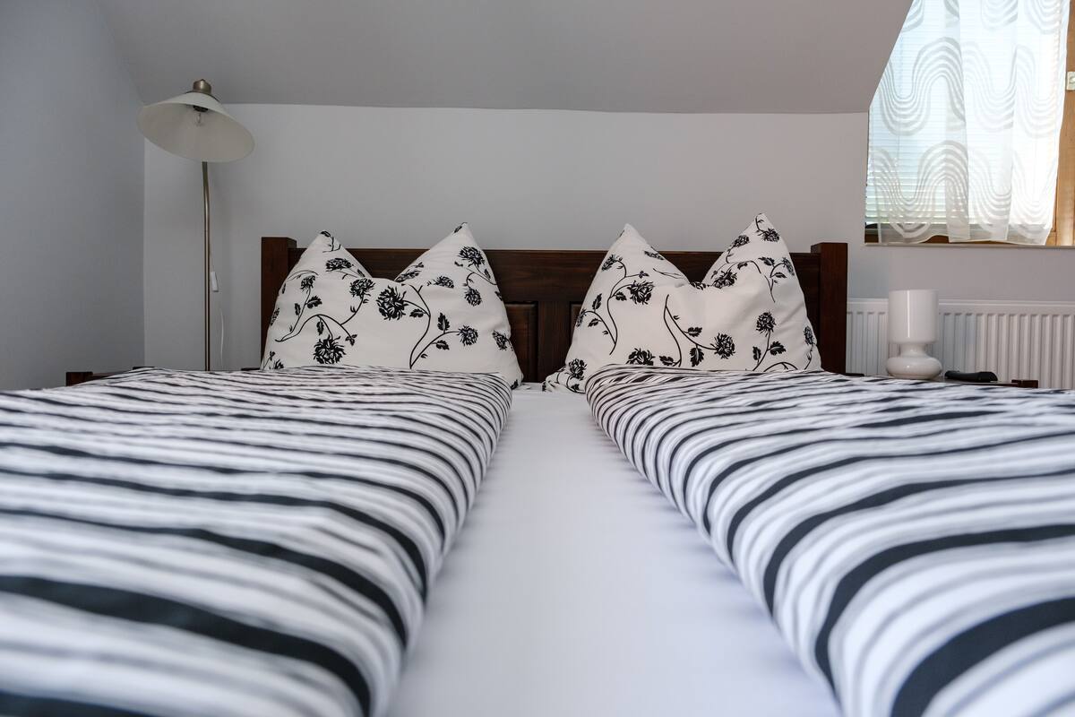 Cama doble con ropa de cama a rayas blancas y negras, dos almohadas florales, una lámpara de noche y una ventana con cortinas translúcidas.