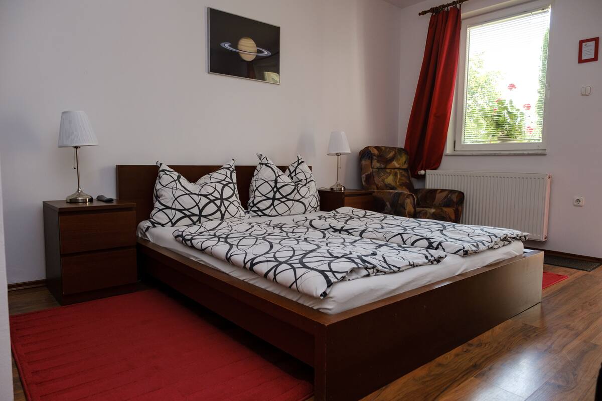 Un dormitorio ordenado con cama, dos mesitas de noche, una silla, una ventana con cortinas y un radiador.