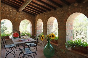 Un patio rústico de piedra con una mesa lista para comer, rodeado de muros de ladrillo en arco y plantas en macetas.