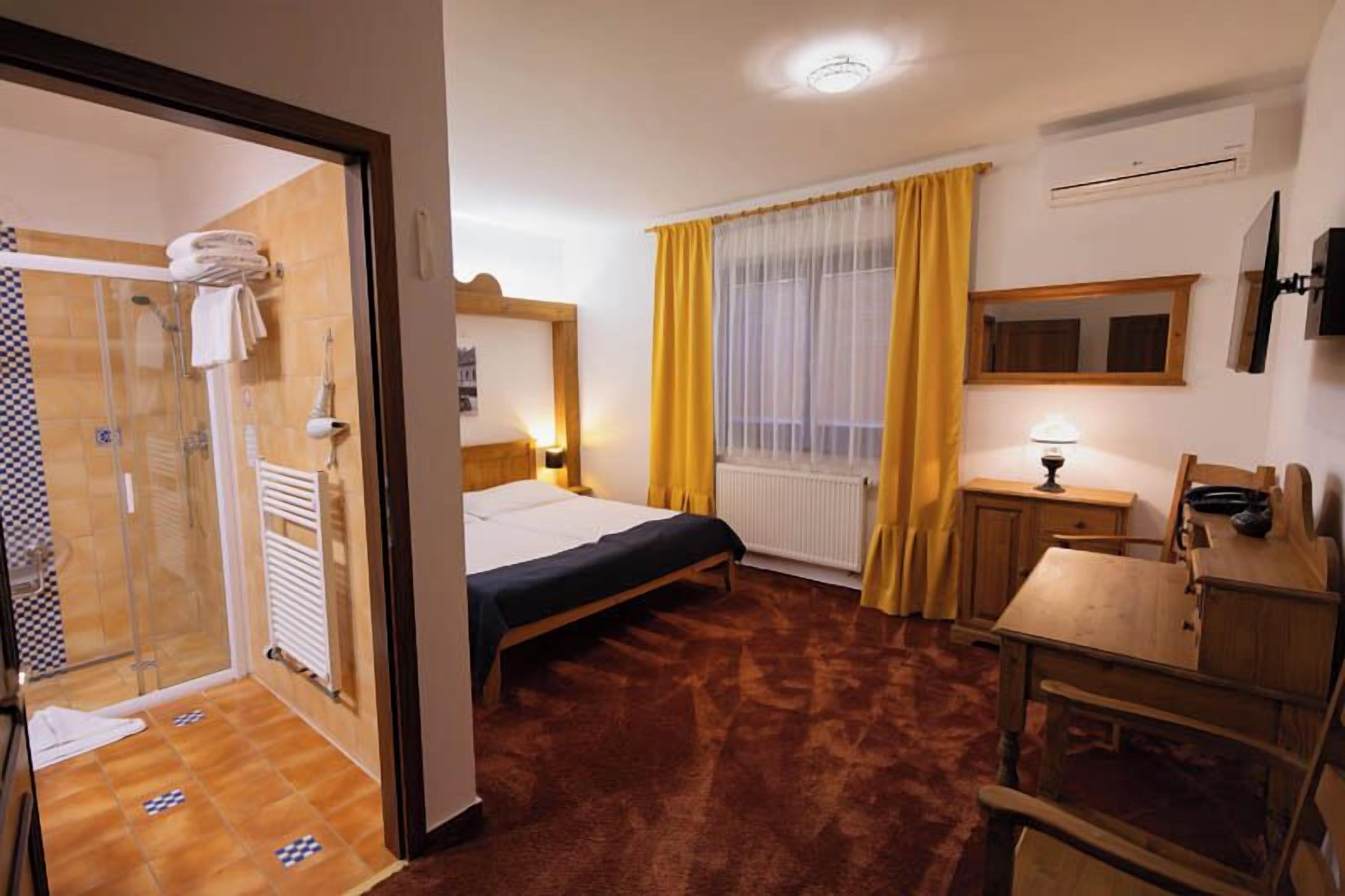 Ett hotellrum med en säng, ett skrivbord, en stol, ett fönster med gula gardiner, ett element och en duschhörna.