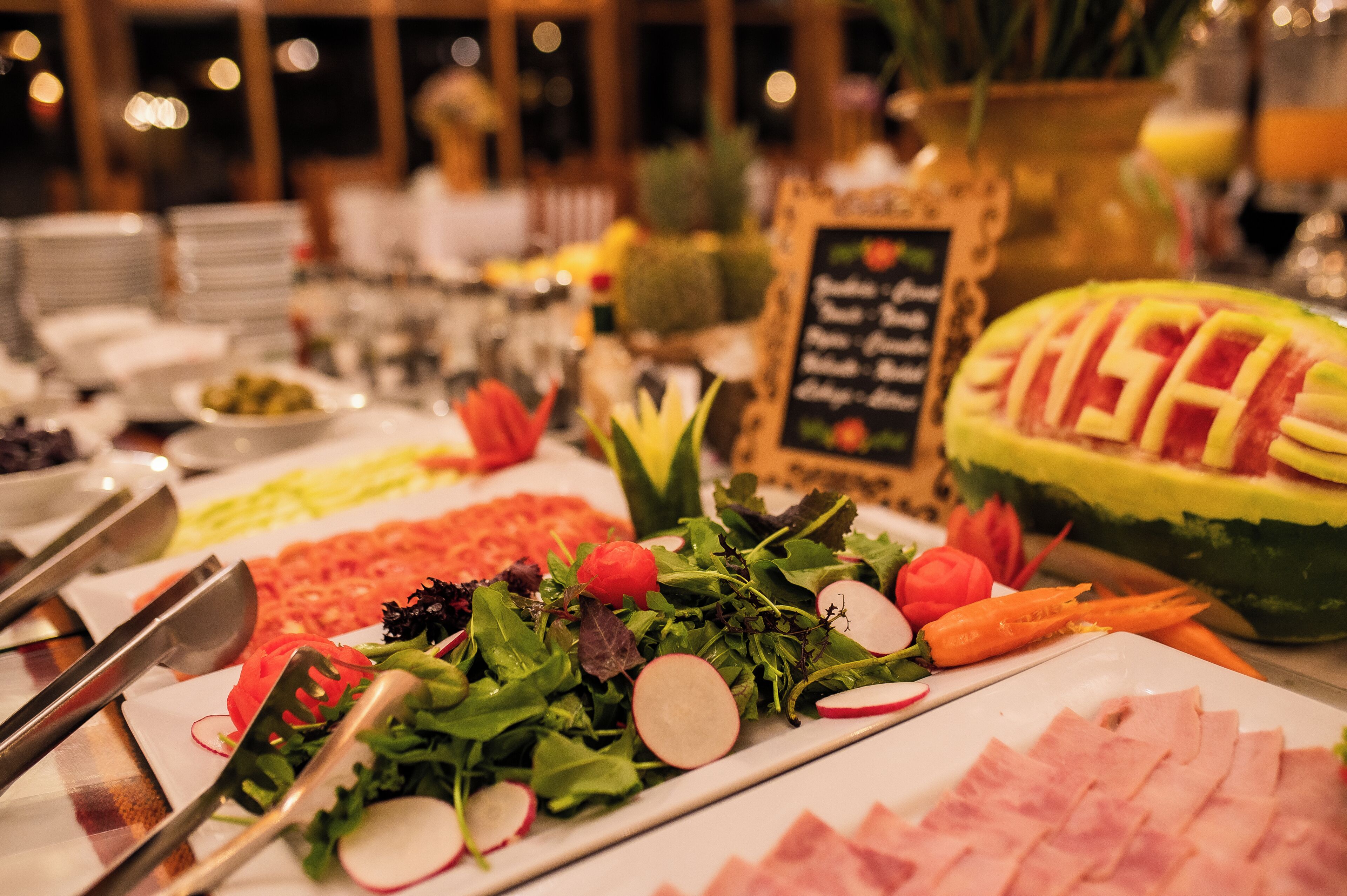 A buffet with various food items including a watermelon carved into a fish shape, salads, and sliced meats.