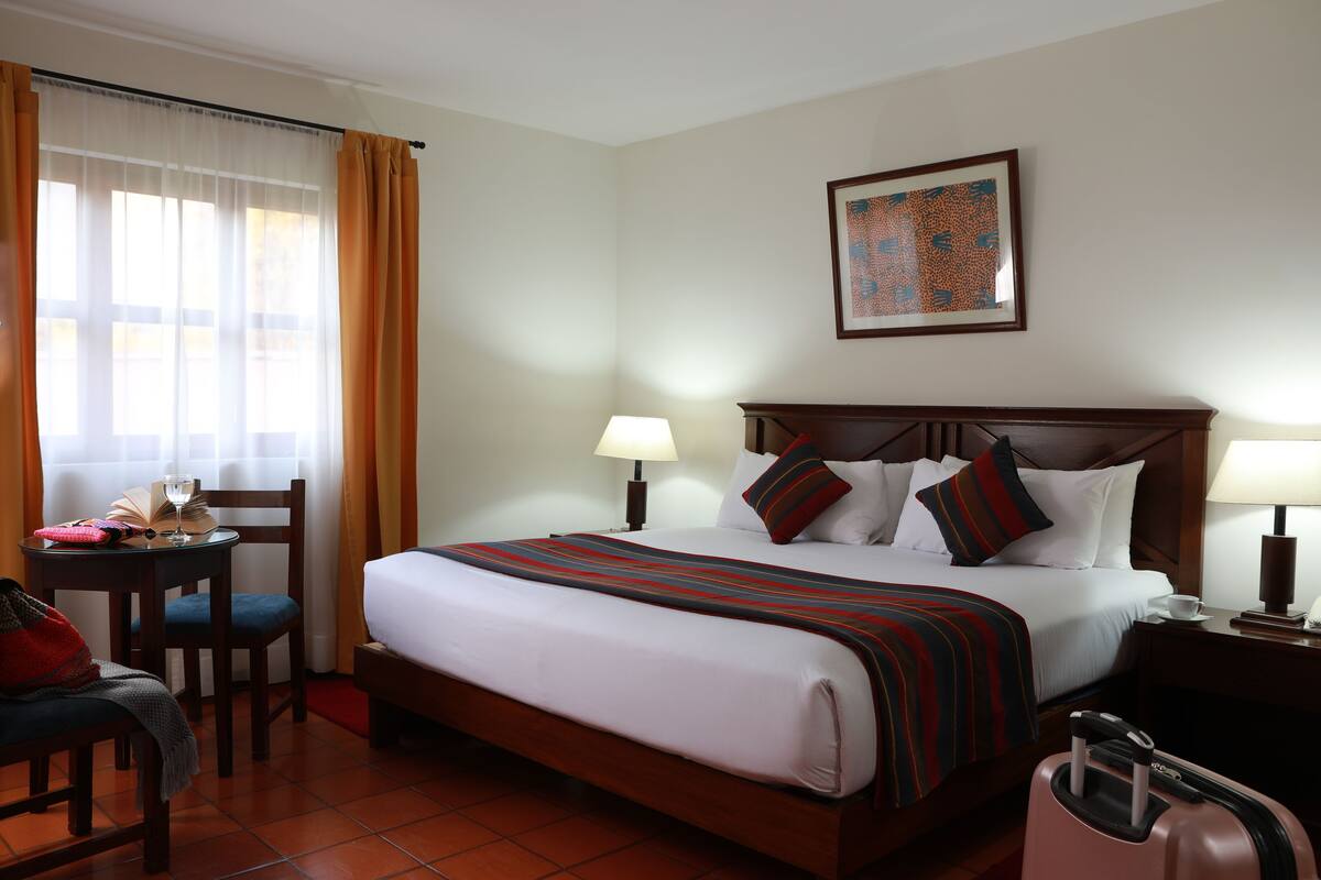 Ein Hotelzimmer mit einem großen Bett, einem Schreibtisch mit Stuhl, einem Fenster mit Vorhängen und einem Gemälde an der Wand.