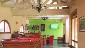 Uma mesa de bilhar em um espaço com parede verde, área de bar e televisão na parede.