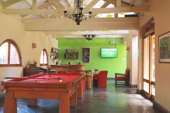 A pool table in a room with a green wall, a bar area, and a television mounted on the wall.