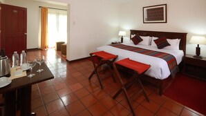 Quarto de hotel com cama, mesas de cabeceira, escrivaninha e uma cadeira, tapete vermelho e uma porta.