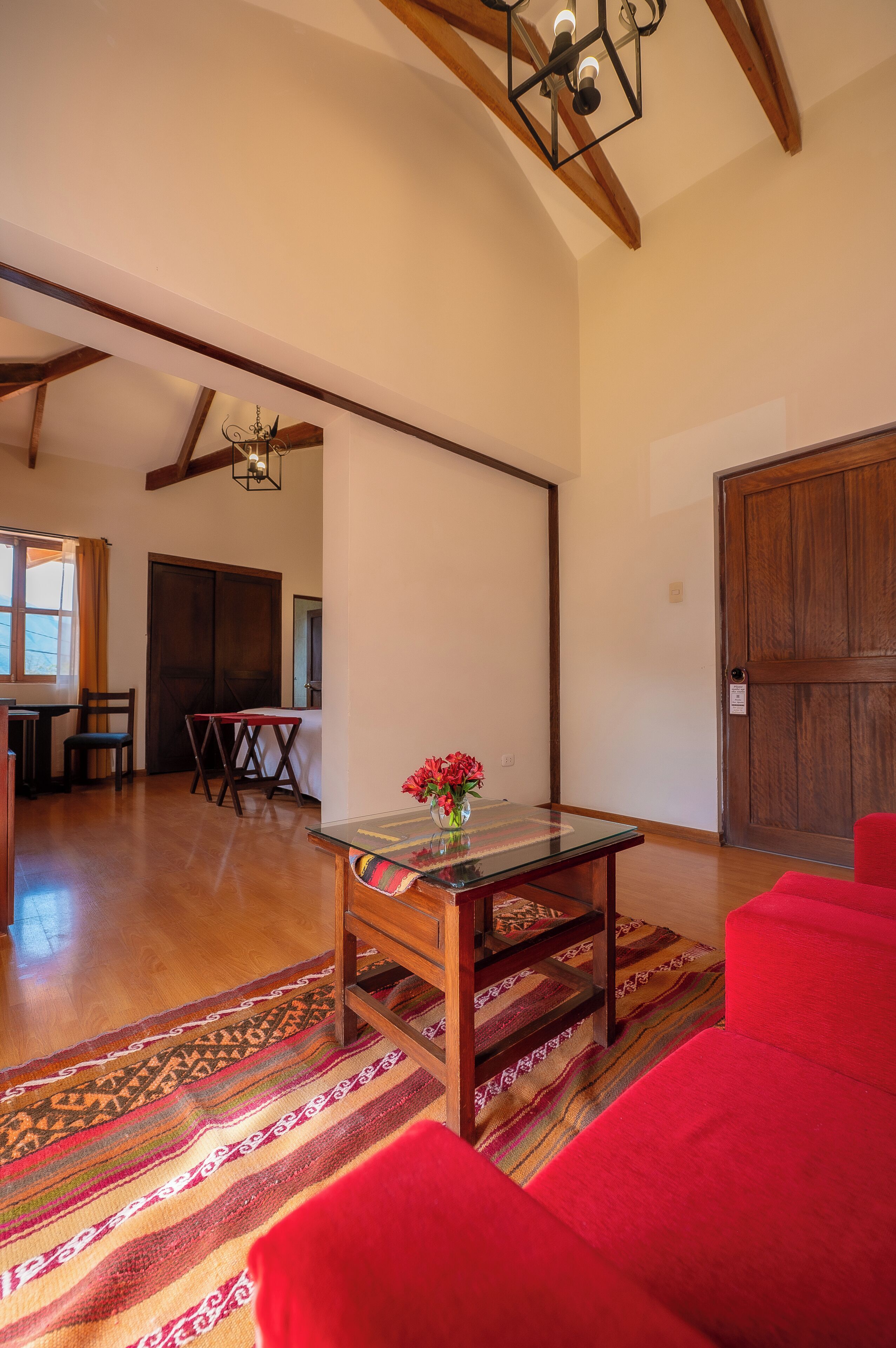 A spacious living room with a wooden floor, a glass coffee table with a vase of flowers, a red sofa, and a wooden door.