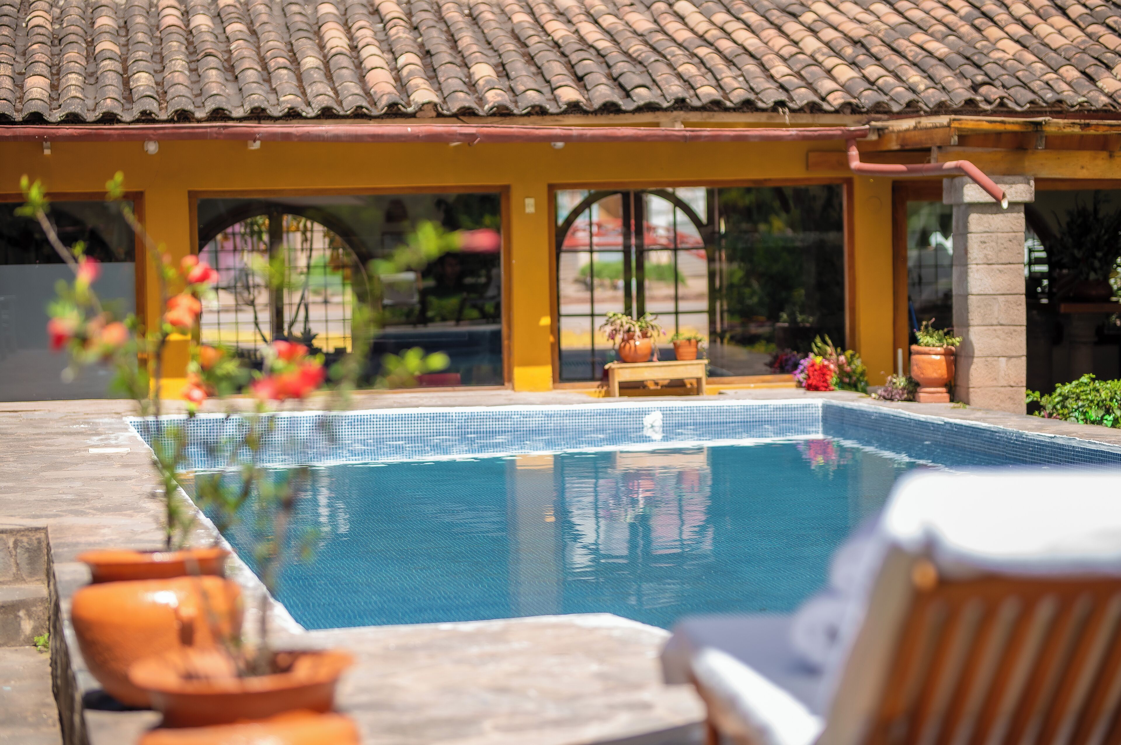 A swimming pool with a tiled deck, a yellow building with a tiled roof, and potted plants.