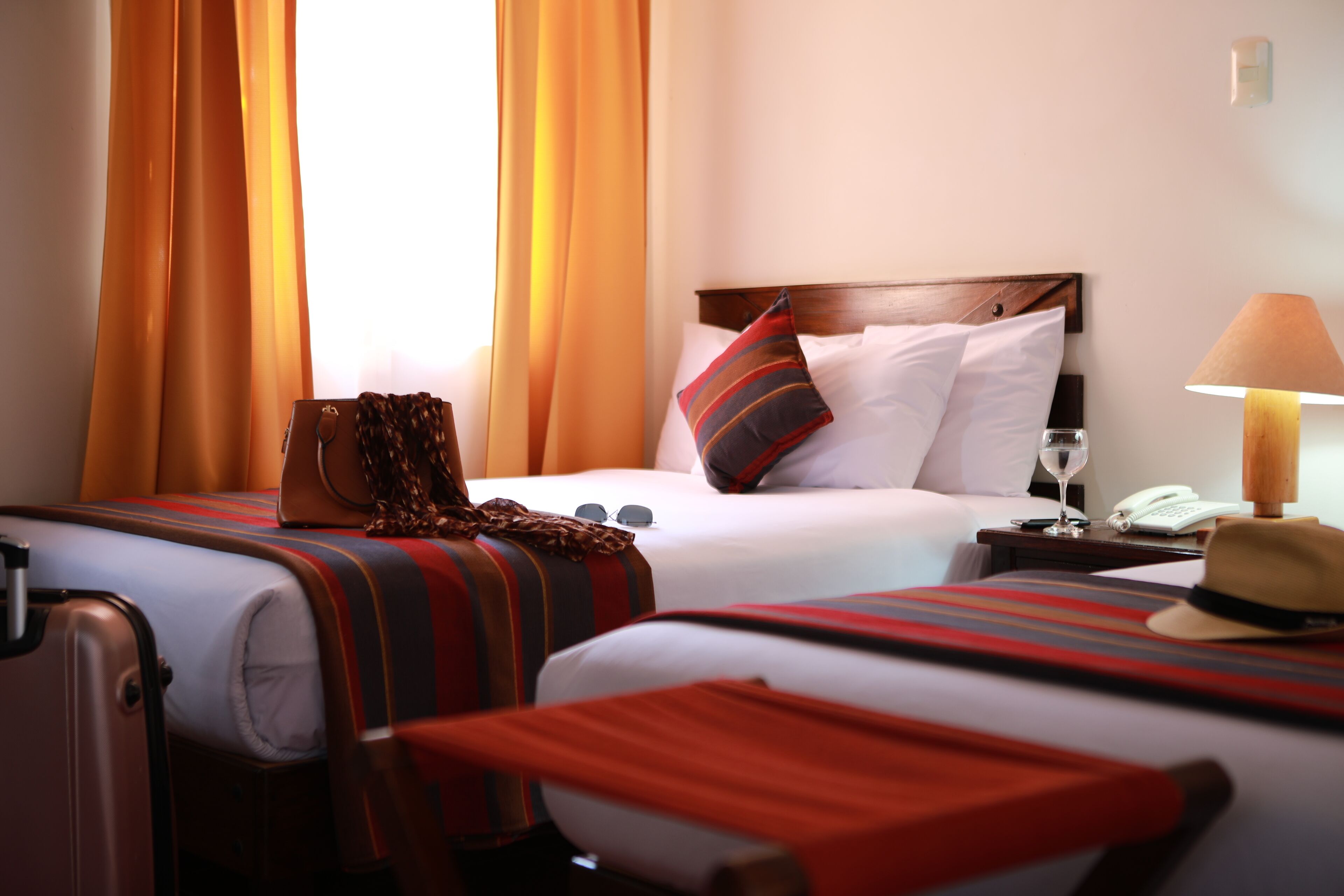 A hotel room with two beds, a wooden headboard, a bedside table with a lamp, a telephone, and a suitcase.