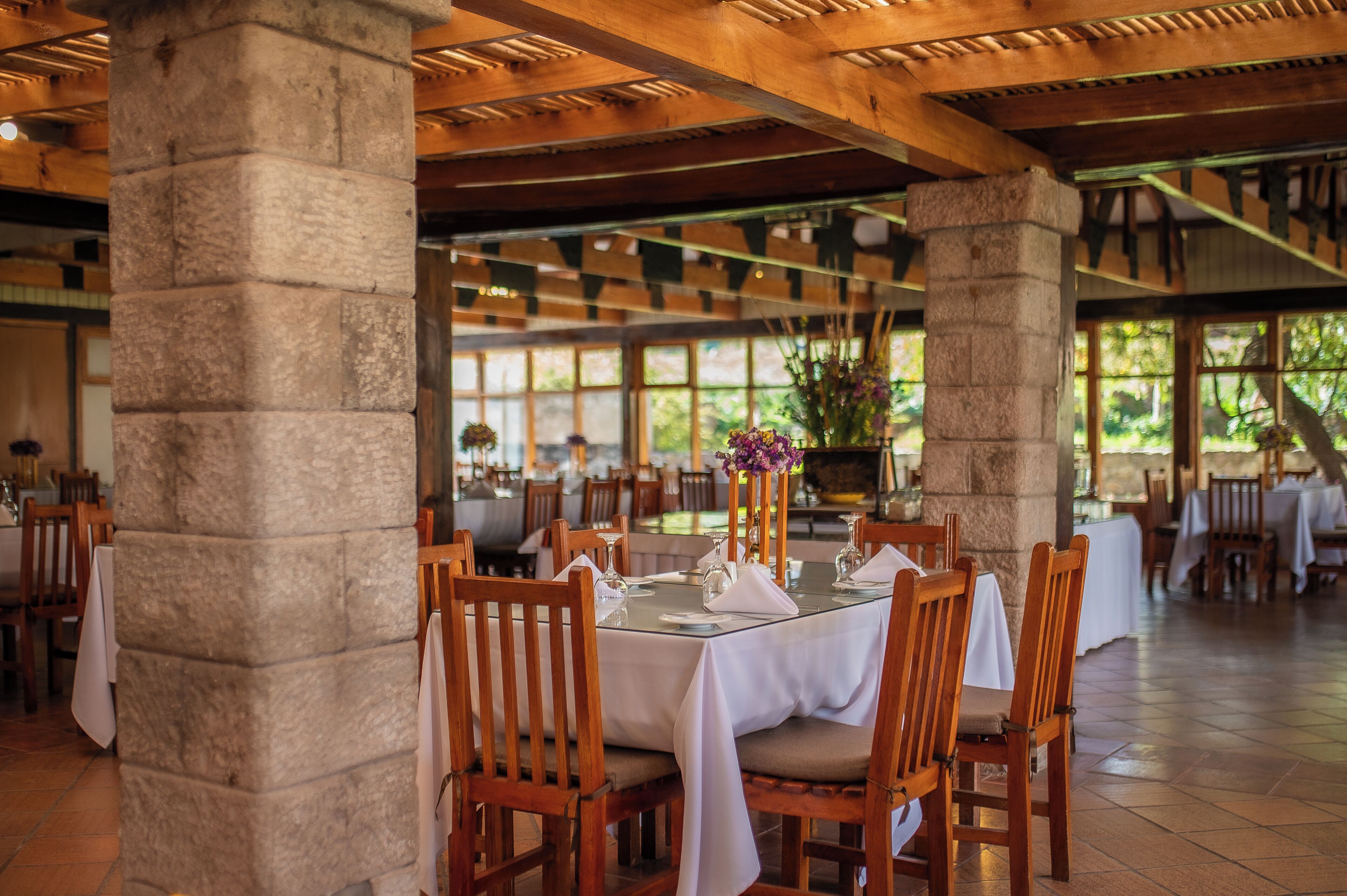 Un restaurante con pilares de piedra, mesas y sillas de madera, y vistas al exterior a través de amplios ventanales.