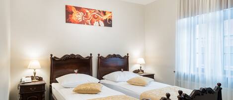 A double bed with a wooden headboard, two bedside tables with lamps, a chandelier, and a large window with sheer curtains.