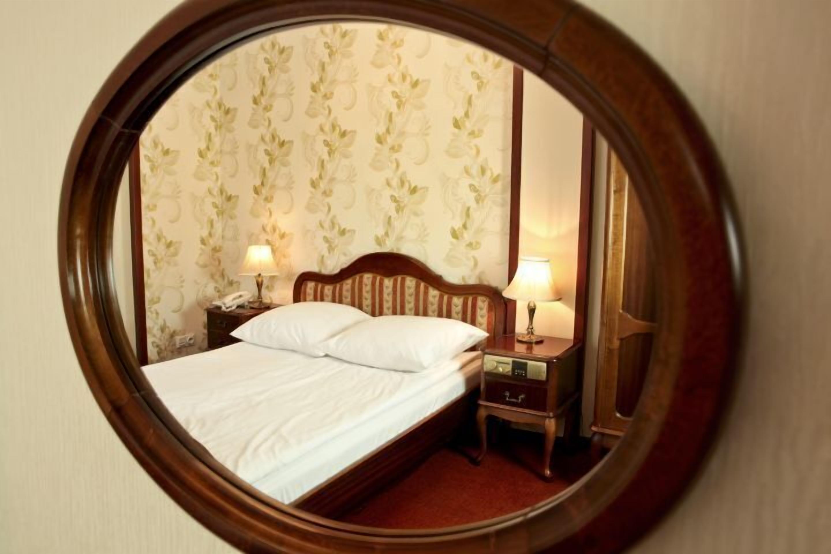 A circular mirror reflecting a neatly made bed with white linens, two bedside lamps, and a wooden nightstand.