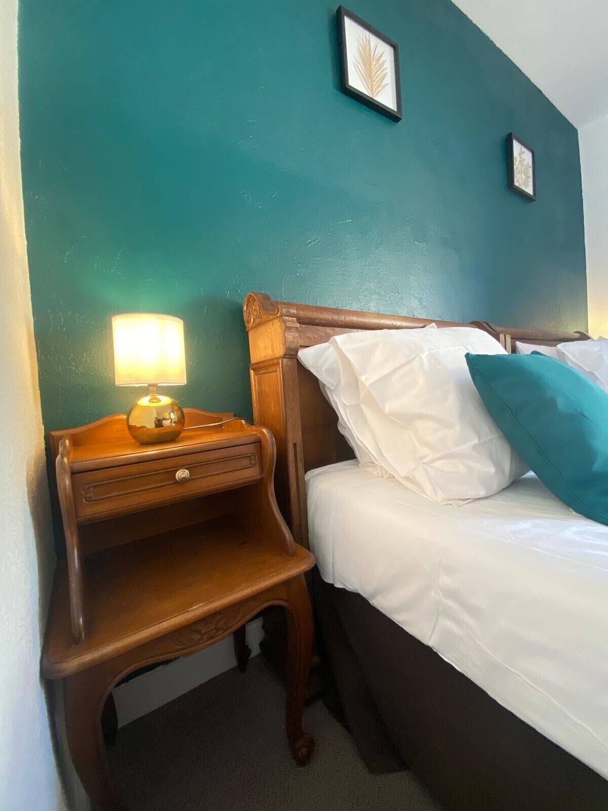 A neatly made bed with white linens and teal pillows, a wooden nightstand with a lamp, and a small drawer. The walls are painted teal, and there are two framed pictures on the wall.