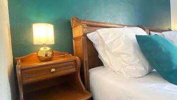A neatly made bed with white linens and teal pillows, a wooden nightstand with a lamp, and a small drawer. The walls are painted teal, and there are two framed pictures on the wall.