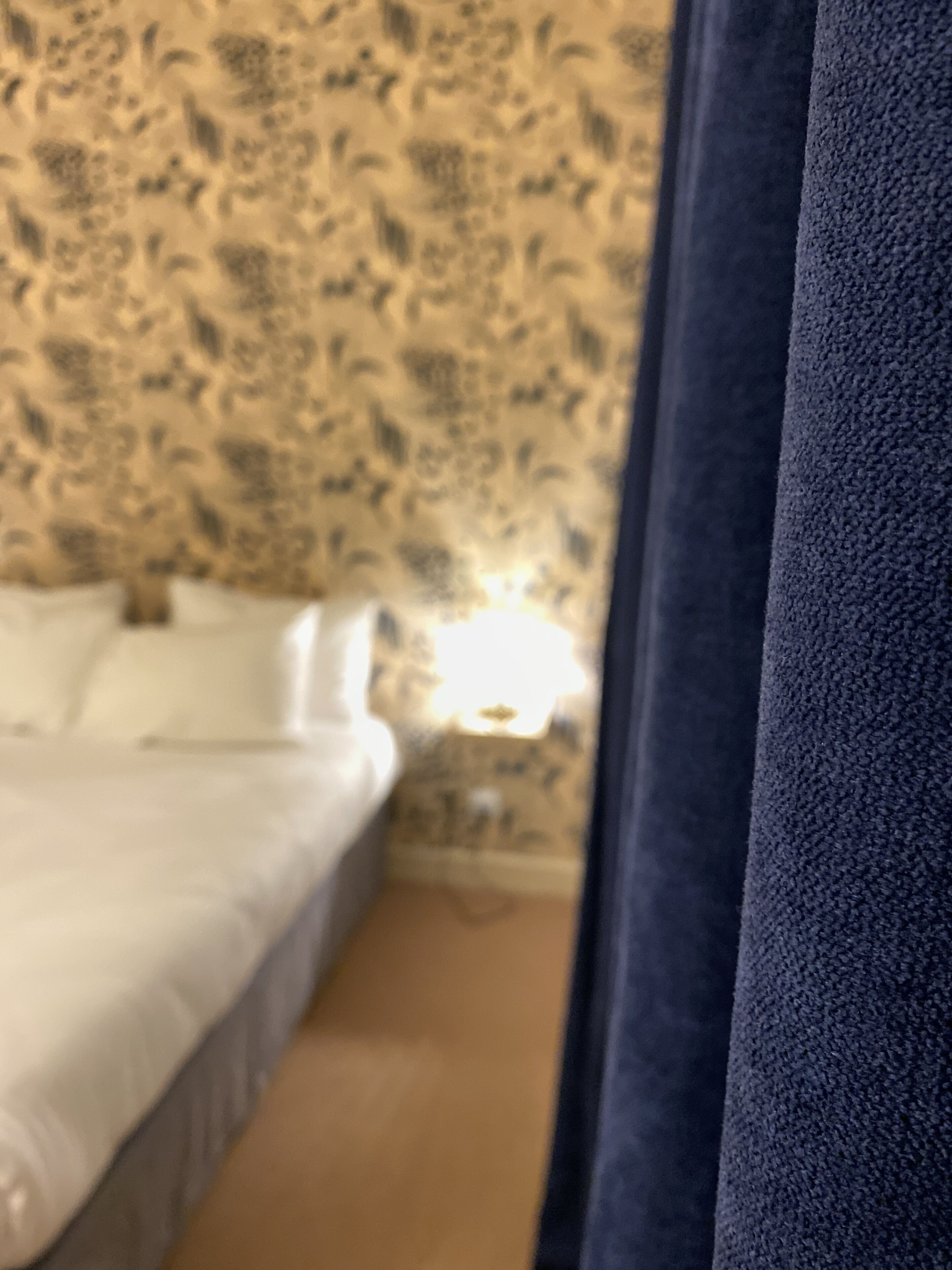 A bed with white linens, a nightstand with a lamp, and a floral-patterned wall.