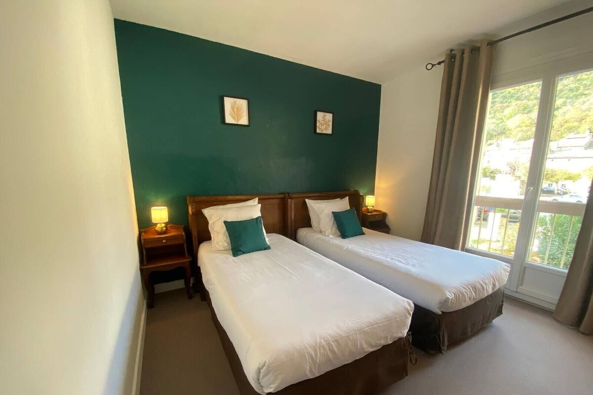 A hotel room with two single beds, a green accent wall, and a view of the outdoors through a large window.