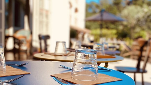 A dining table with a clear glass and a napkin, set outdoors with a building and trees in the background. - Logis Hôtel Médiéval, Montélimar Nord (Rochemaure)