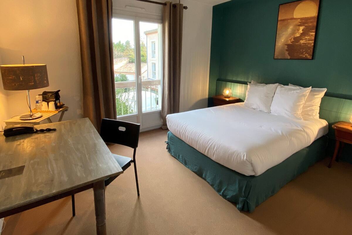 A hotel room with a bed, a desk, a chair, a lamp, a painting, and a view of the outdoors.