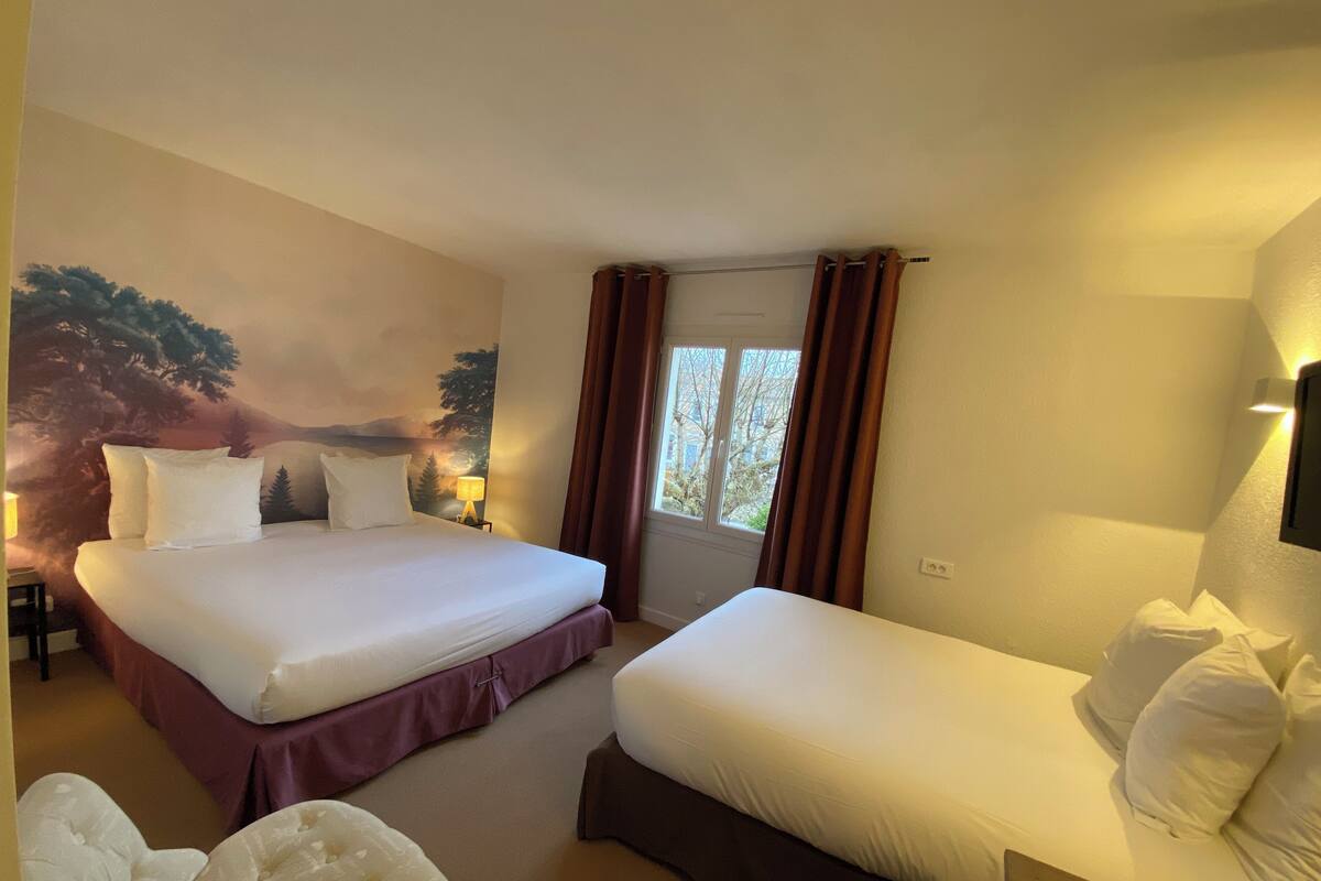A hotel room with two beds, a large window with curtains, a wall mural, and a bedside table with a lamp.