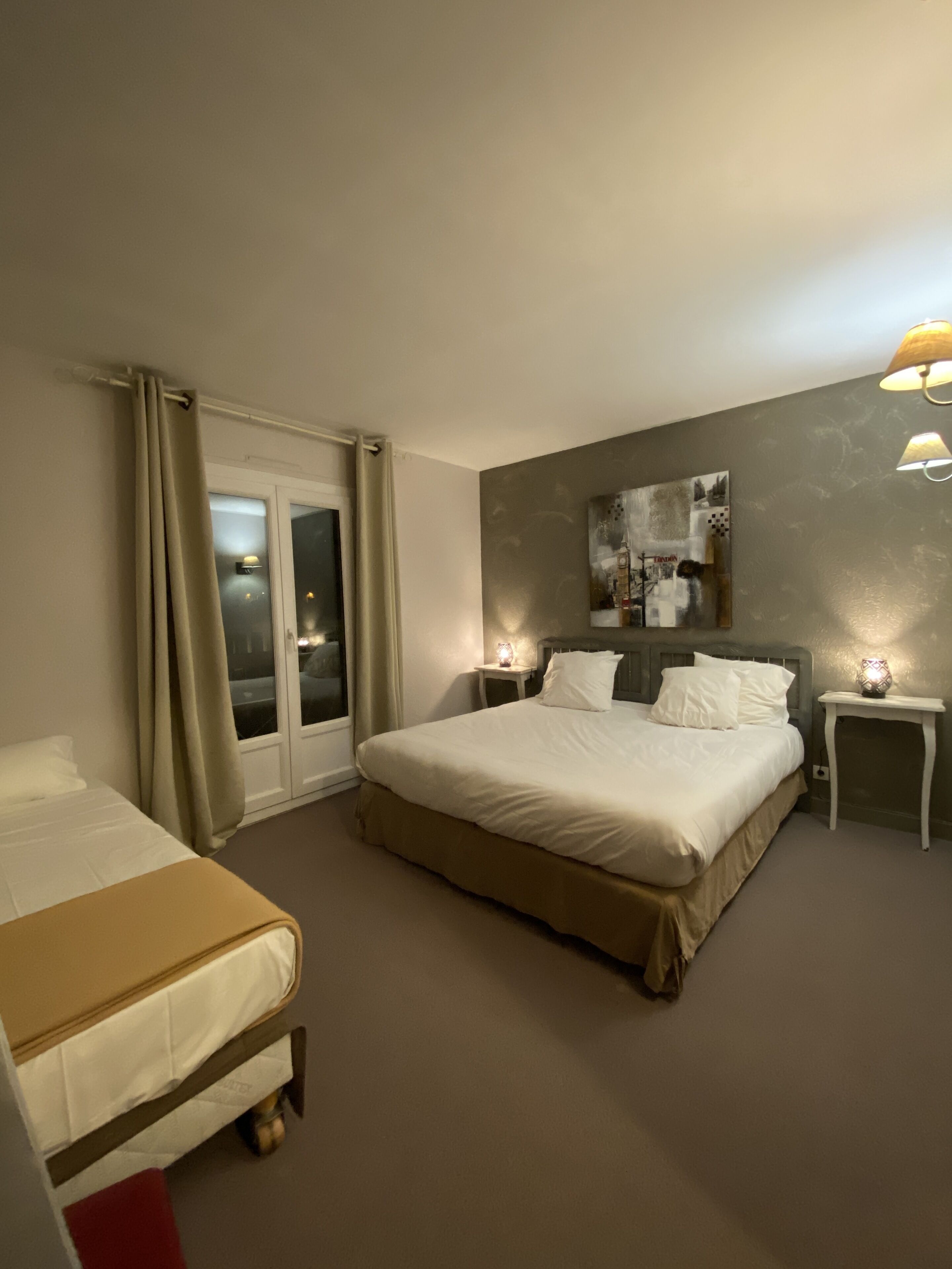 A hotel room with a large bed, a smaller bed, a nightstand with a lamp, a wall-mounted artwork, and a window with curtains.