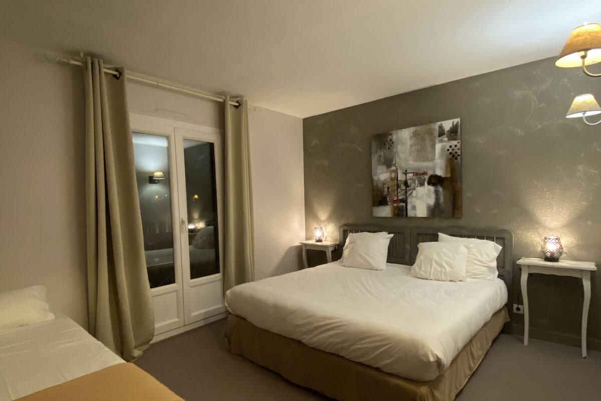 A hotel room with a large bed, a smaller bed, a window with curtains, a nightstand, and a wall-mounted lamp.