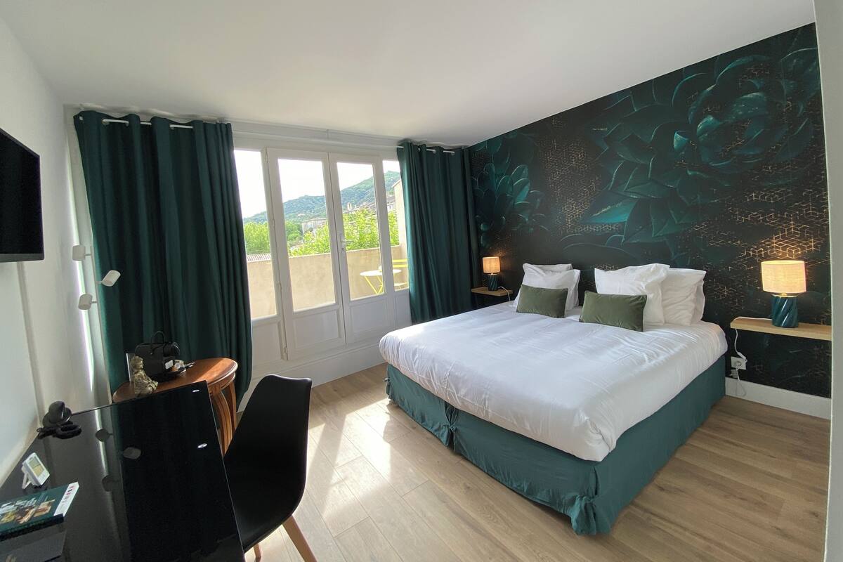 A hotel room with a large bed, a desk with a chair, a TV, and a view of greenery through the windows.