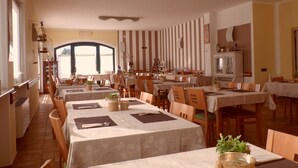 A dining area with multiple tables and chairs, a kitchen area, and a large window providing natural light. - Hotel Agli Ulivi (Valeggio sul Mincio)