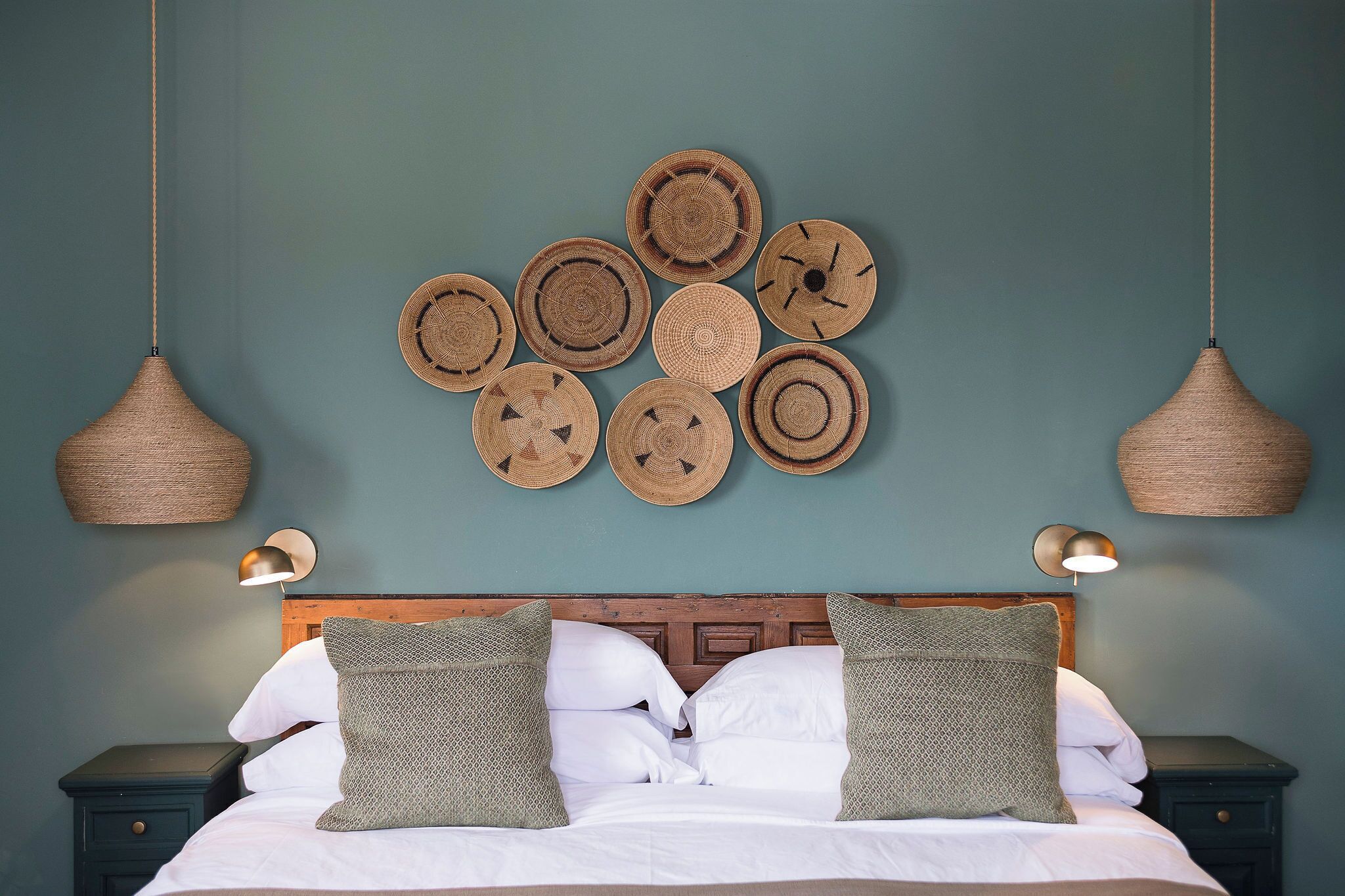 A neatly made bed with white linens and two green pillows, a wooden headboard, and a wall adorned with woven circular wall hangings.