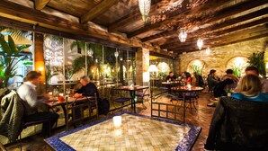 An indoor dining area with wooden beams, stone walls, and hanging lanterns. Tables are arranged around a central tiled area with a candle on it. - La Casa del Califa (Vejer de la Frontera)