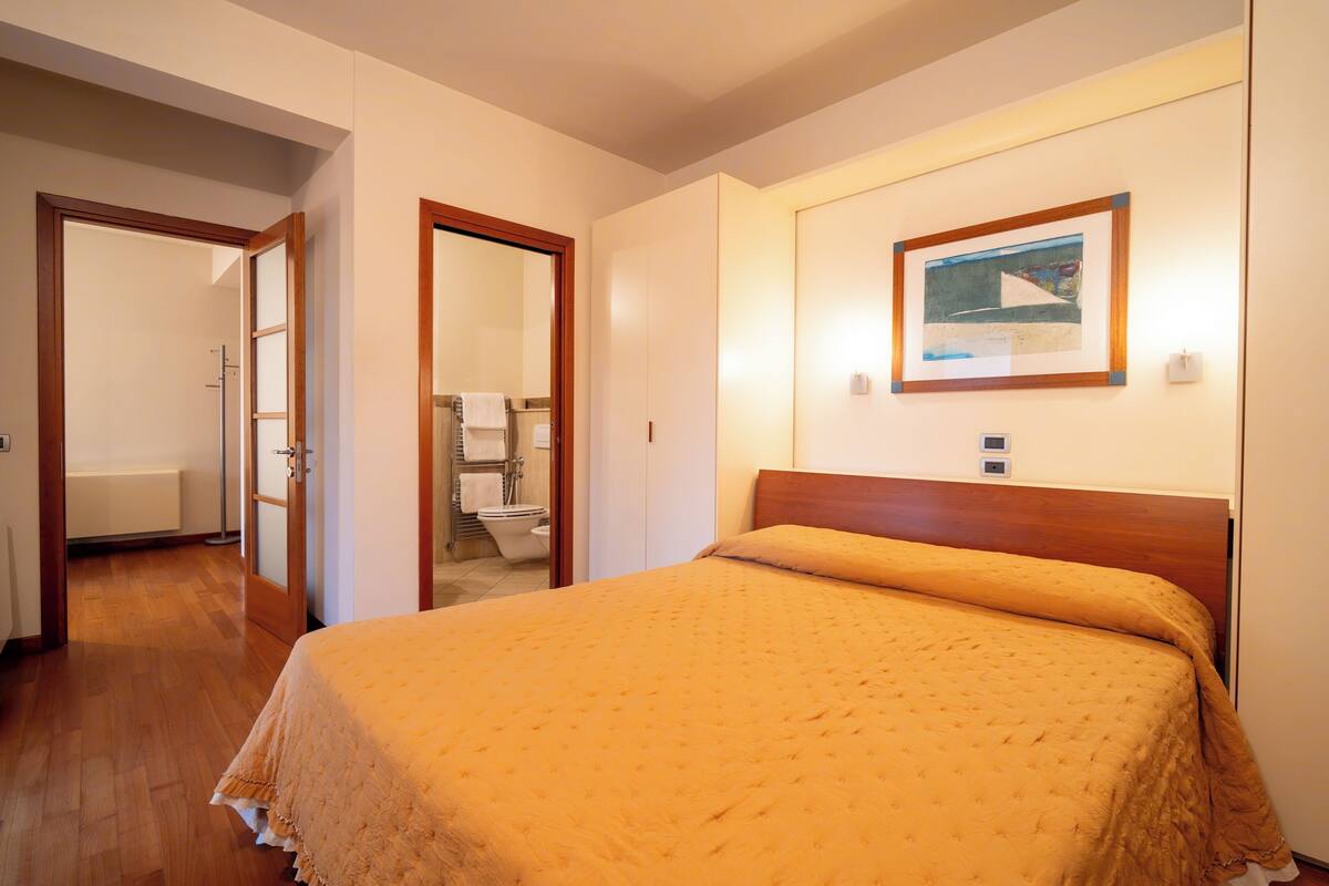 Una camera d'albergo con un letto di legno, un quadro appeso alla parete e un bagno visibile attraverso una porta aperta.
