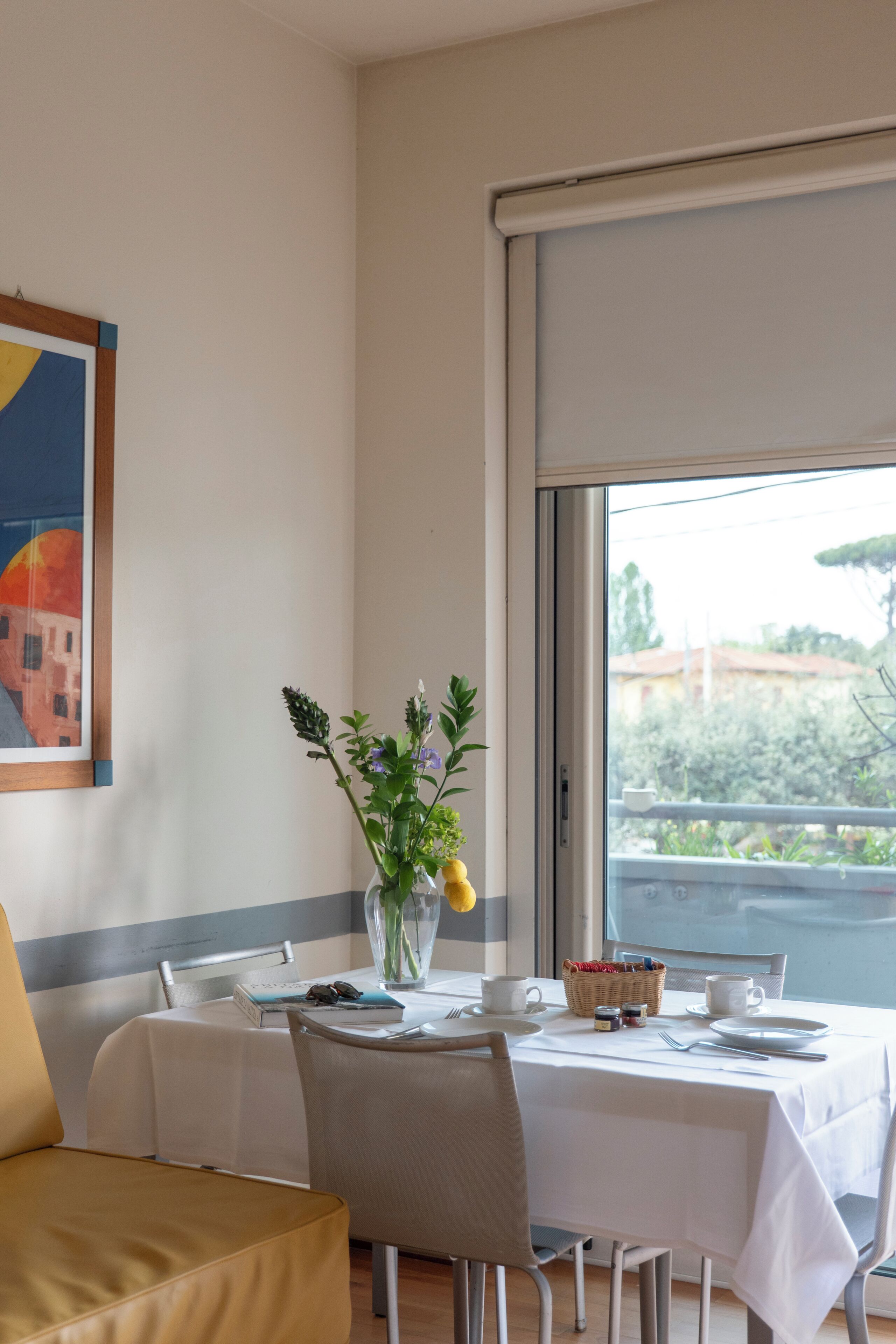 A dining area with a table set for two, a vase with flowers, and a painting on the wall.