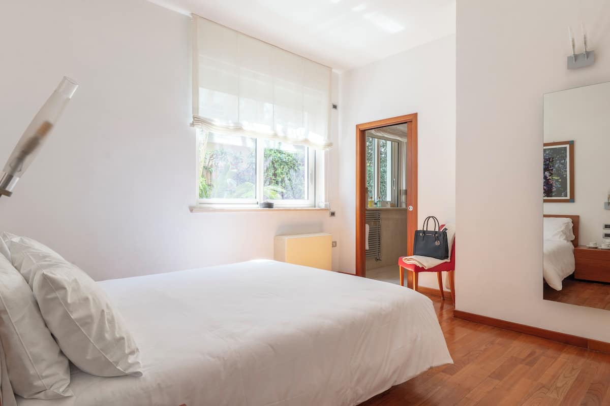 Una camera da letto con un letto, una finestra con tende leggere, una sedia, un comodino e uno specchio.