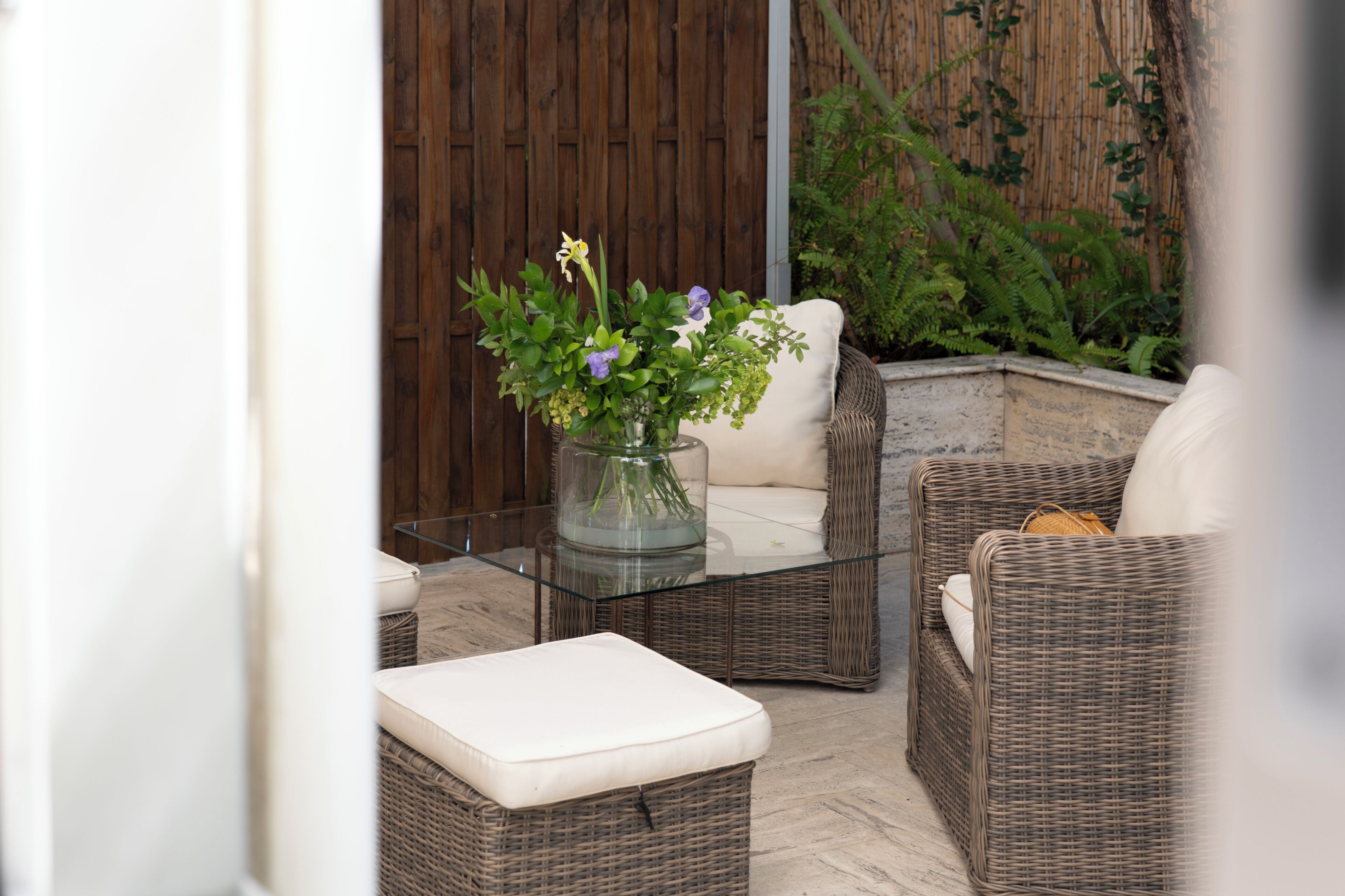 Un área de estar al aire libre con muebles de mimbre, una mesa de vidrio, cojines y un florero con flores.