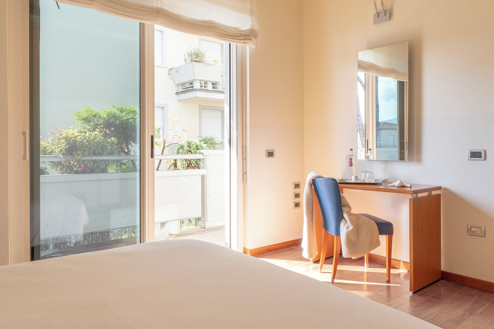 Un dormitorio con cama, escritorio, silla, espejo y puerta corrediza de vidrio que da a un balcón.