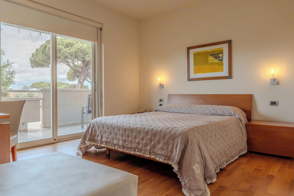 Camera da letto con un letto grande, un comodino in legno, un tavolino da notte, una sedia e una porta scorrevole in vetro che dà su una zona esterna con vista su alberi e montagne.
