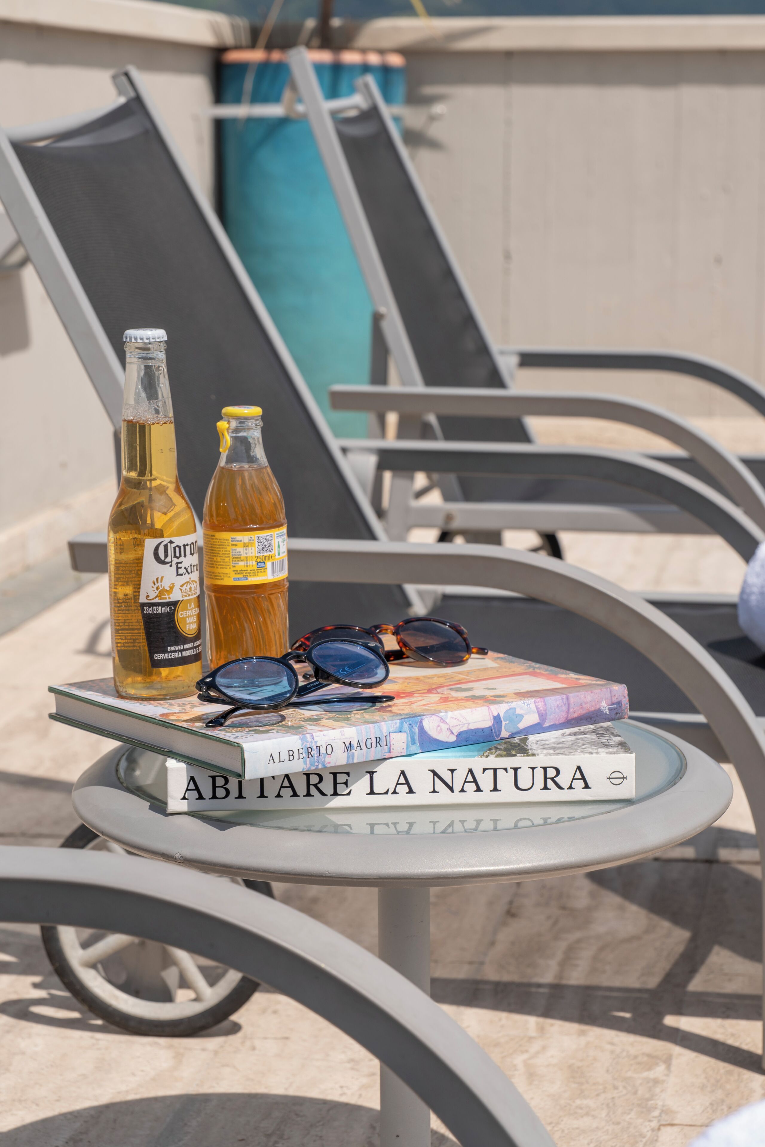 A table with a book titled 'ABITARE LA NATURA' and two bottles of Corona beer, sunglasses, and a metal chair.