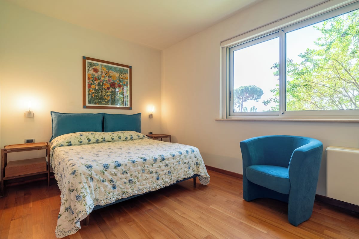 Una camera da letto con un letto, una poltrona blu, un comodino, una lampada e una finestra vista alberi.