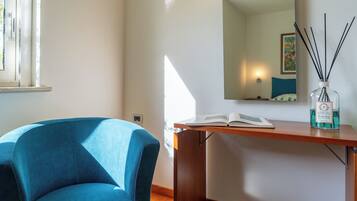 A room with a blue armchair, a wooden desk with a book and a bottle of reed diffuser, and a mirror on the wall.